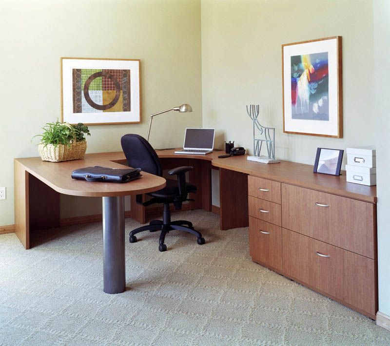 An office with a wooden L-shaped desk, computer, black chair, and storage cabinets. Artwork hangs on the walls.
