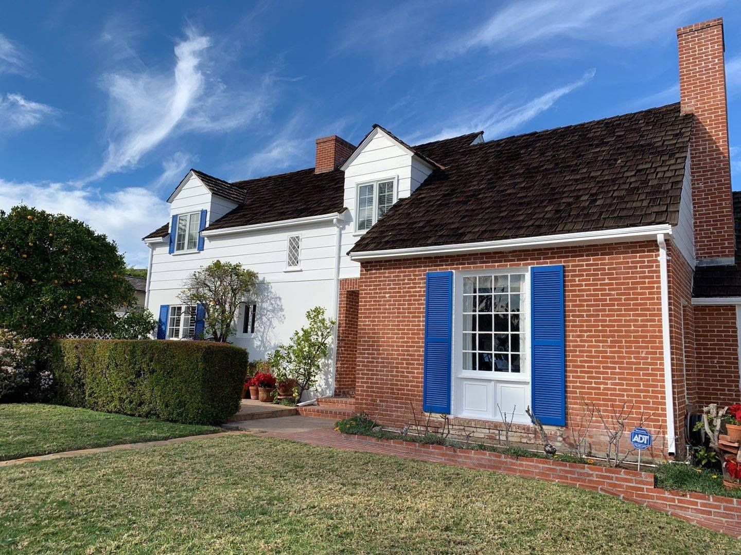 White house with red brick wing, blue shutters, brown roof, and brick chimney on a green lawn under a blue sky.