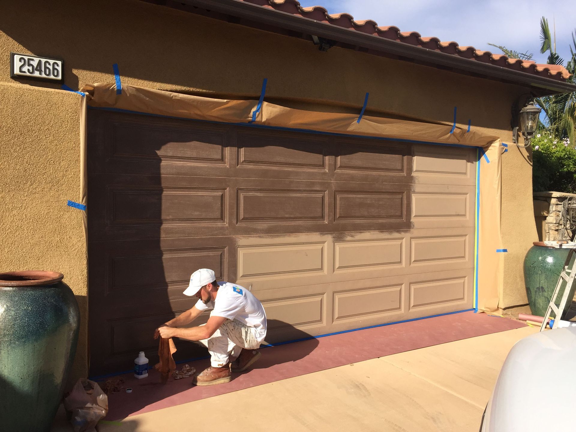 Person painting a brown garage door on a stucco building; masking tape and paper protect surrounding areas.