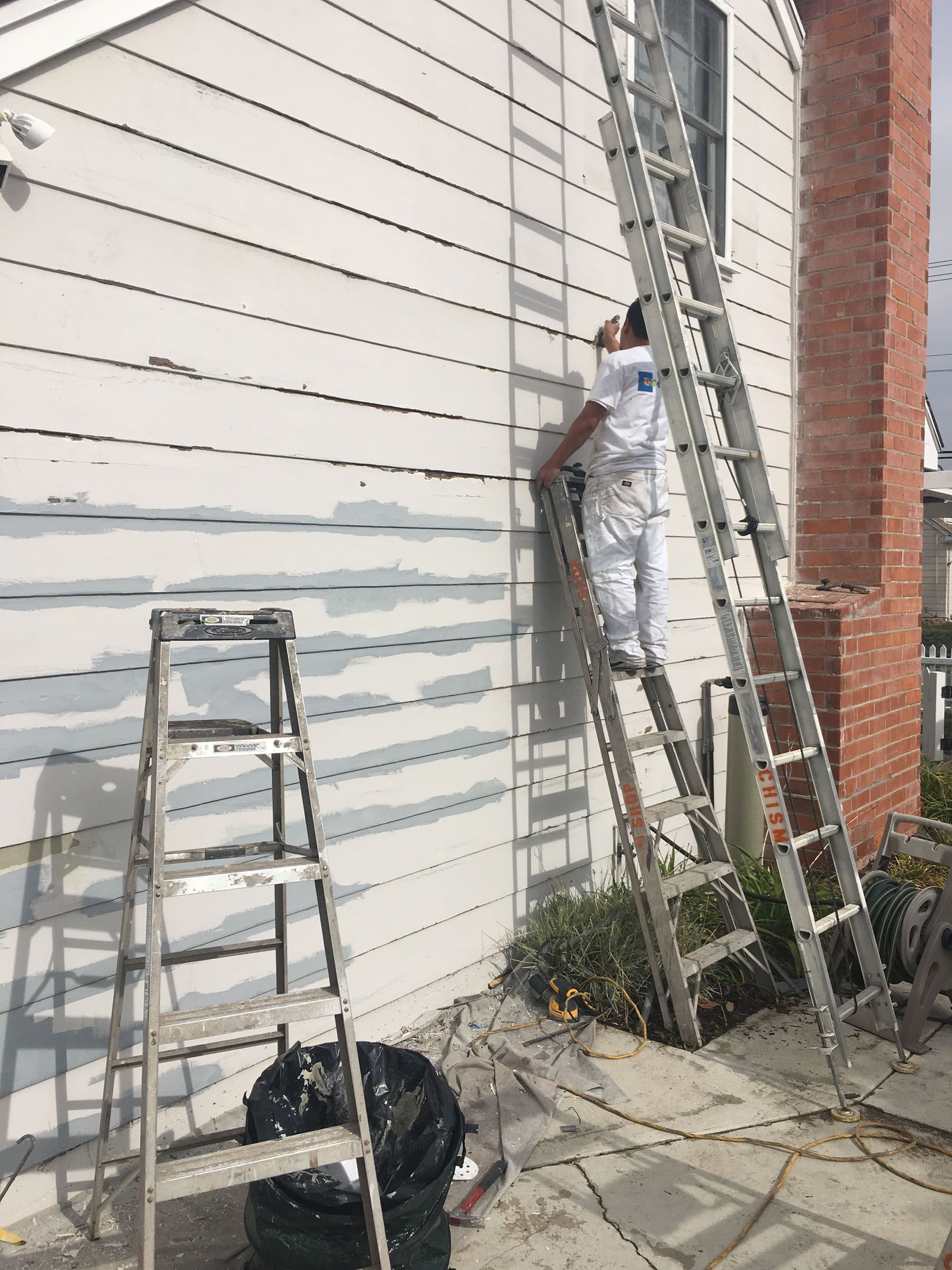 Person painting siding of a house, using a ladder. A second ladder is nearby.