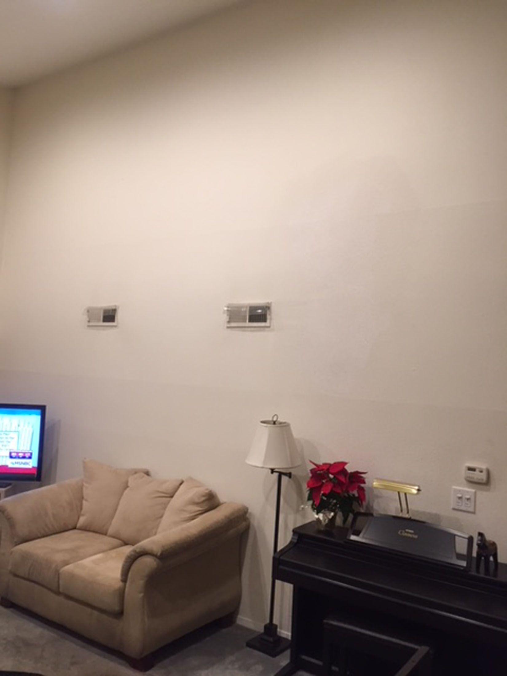 Beige sofa, piano, and poinsettias against a tall, off-white wall with two vents.