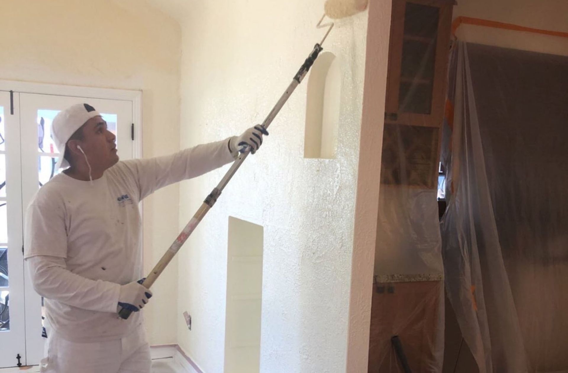 Person painting an interior wall with a paint roller on a long handle.