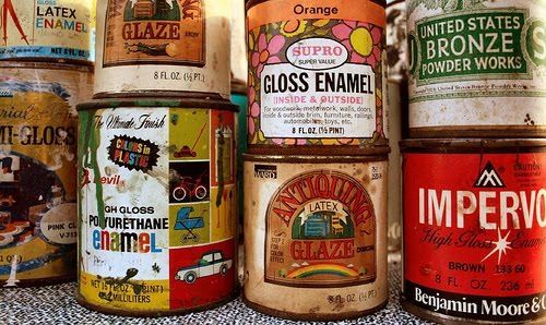 Close-up of vintage paint cans in various colors and designs, stacked on a shelf.