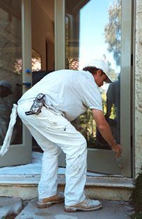 Painter in white overalls and hat working on a glass door frame outdoors.