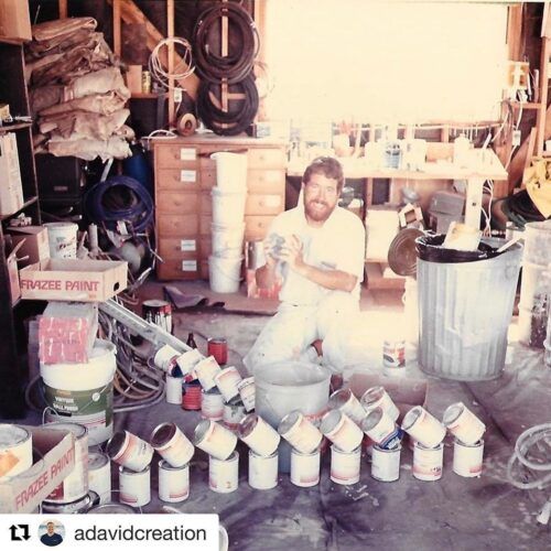 Man in paint-covered clothes kneels amid paint cans in a cluttered workshop.