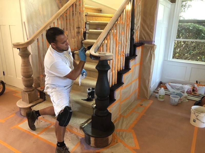 Man painting a black staircase banister, wearing knee pads, in a room covered with protective paper.