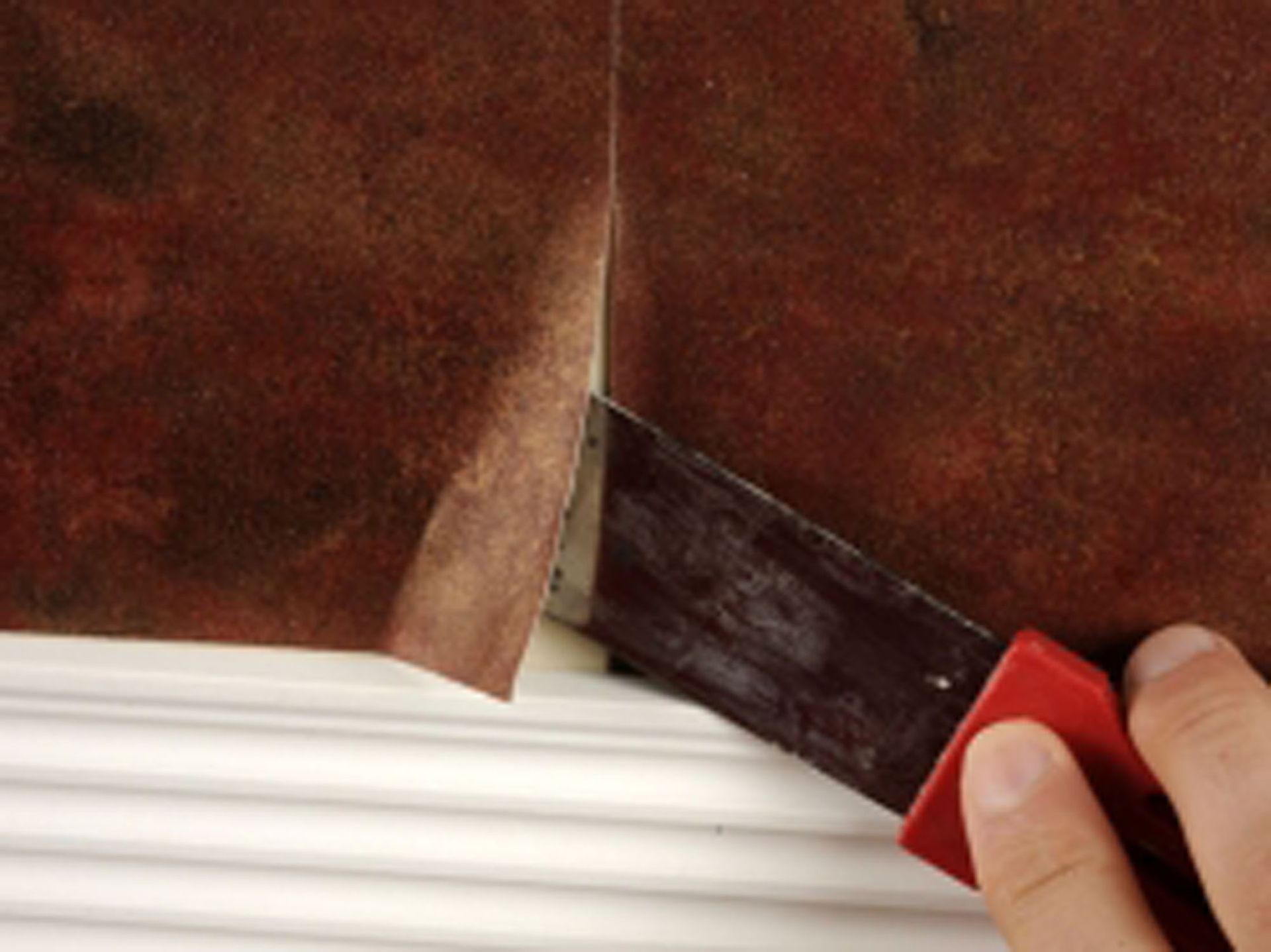 A person's hand using a utility knife to trim wallpaper at the edge of a wall near a white molding.