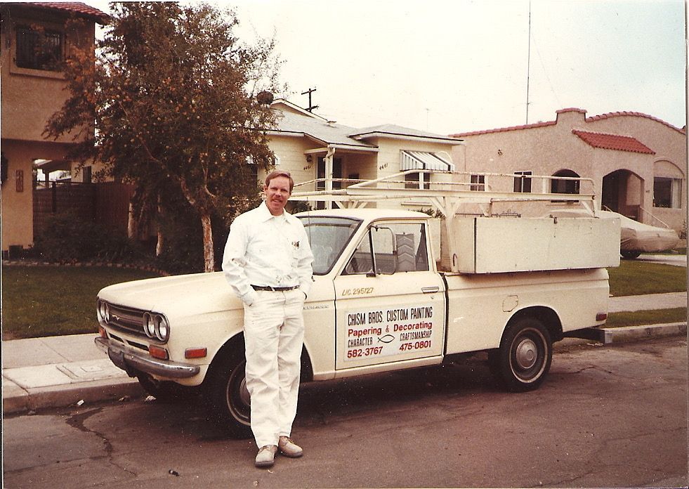 Man in white overalls stands next to a white pickup truck with 