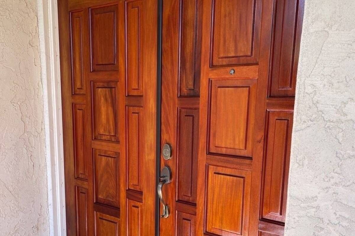 Wooden double doors, polished brown, with rectangular panels. A handle and lock are visible. Set in a stucco wall.