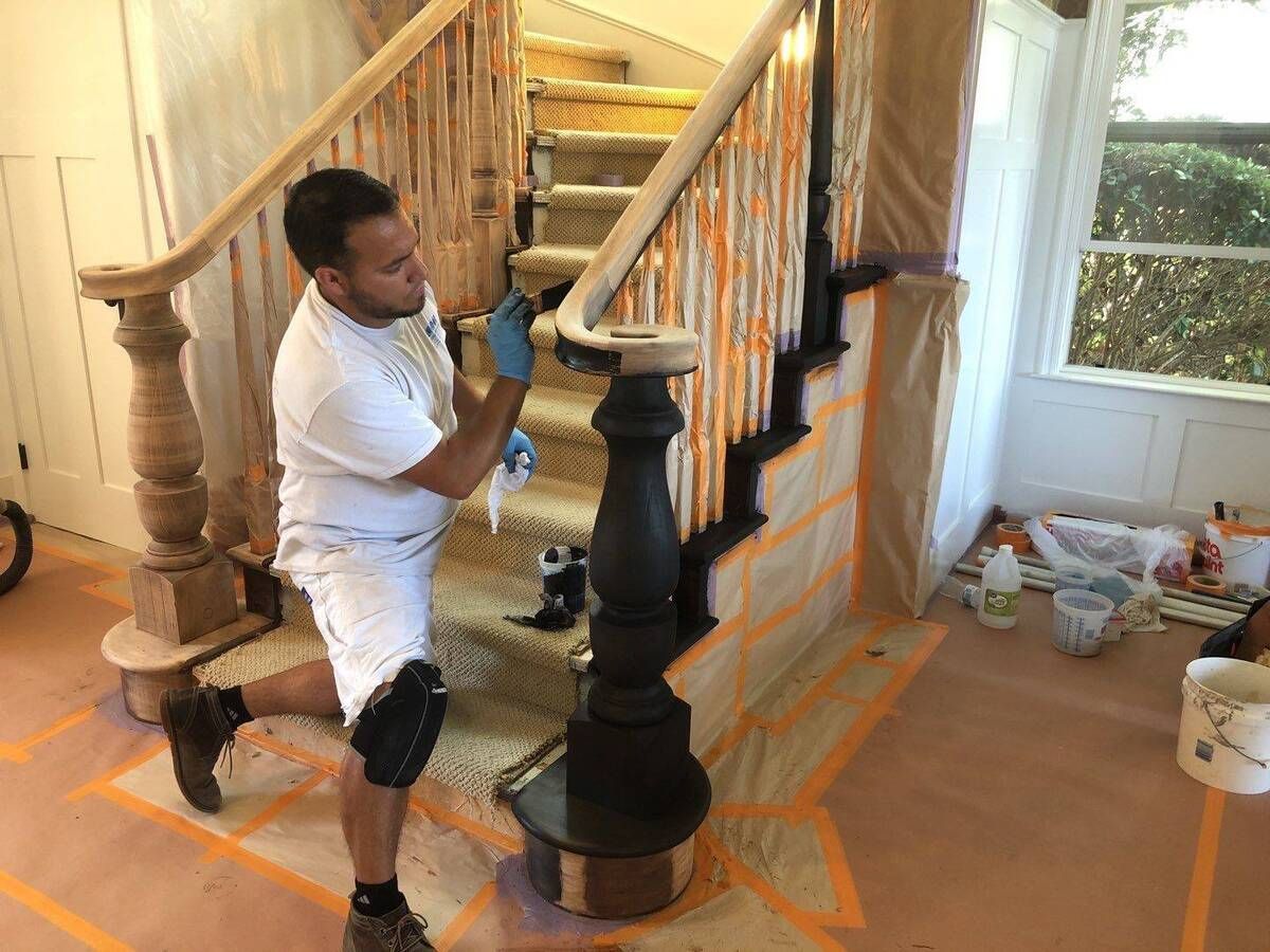 Person paints a stair railing in a home, kneeling with knee pads. Brown and black wood, with masking and drop cloths.
