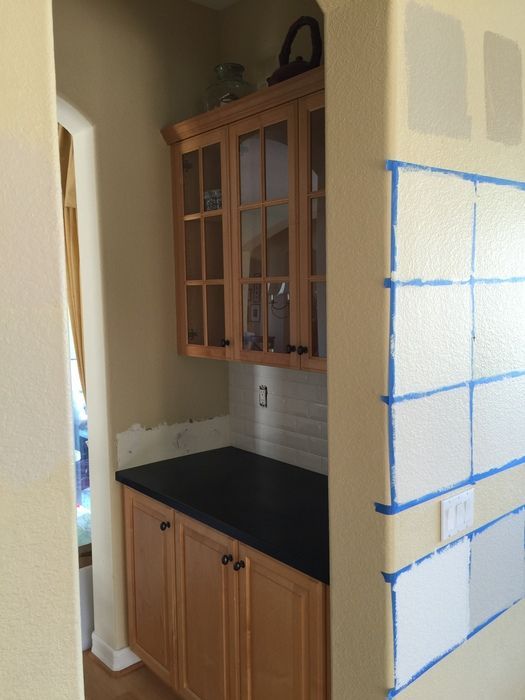 A built-in kitchen cabinet area with light wooden cabinets, black countertop, and off-white tiled backsplash.