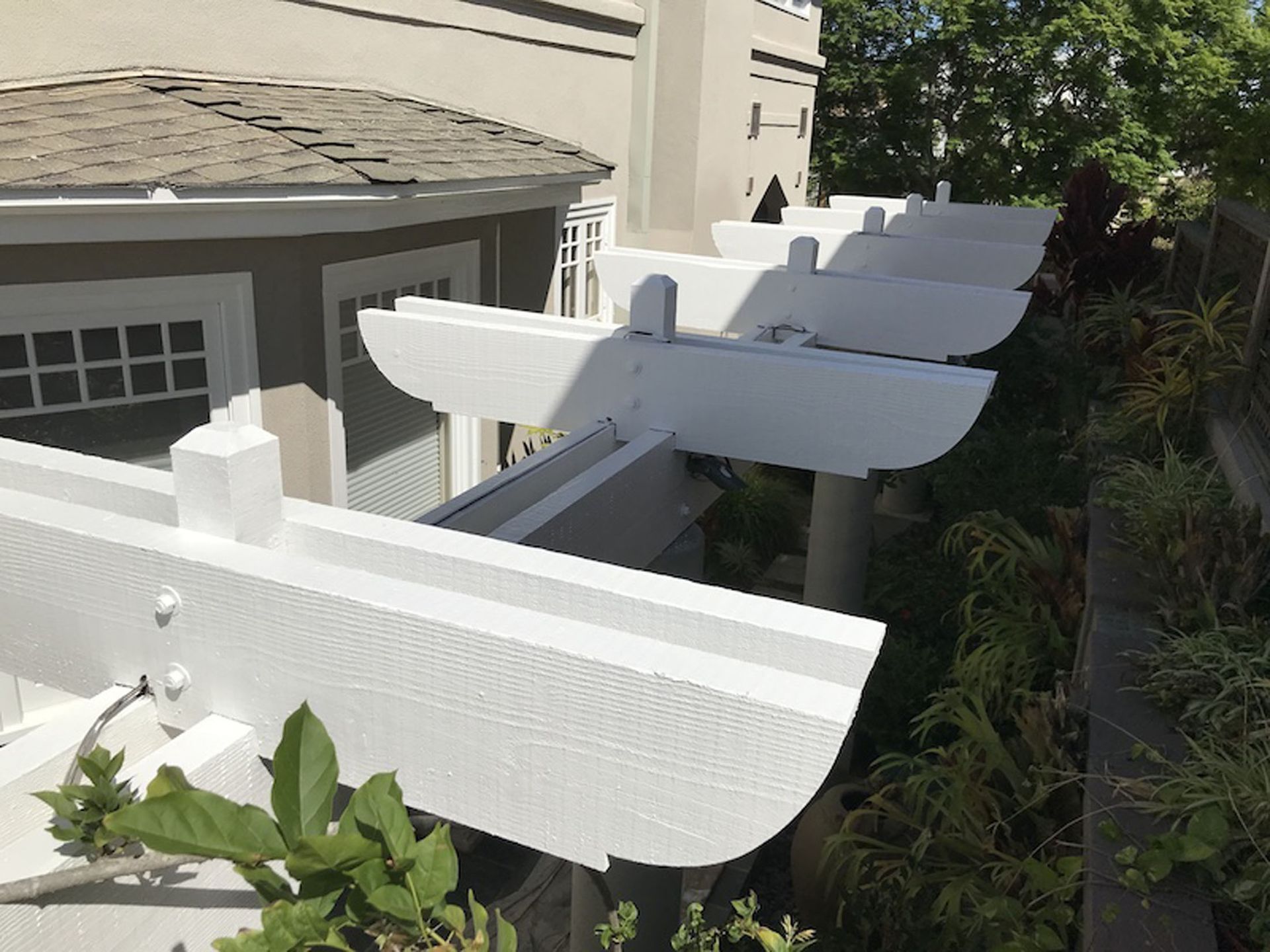 White pergola extending from a light-colored house, with surrounding greenery.