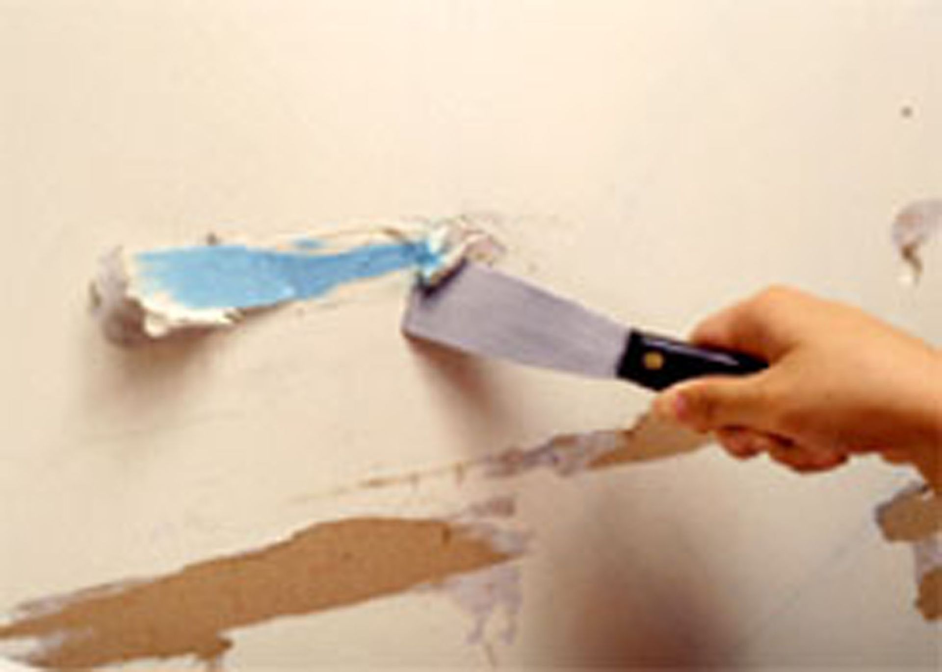 Hand holding a putty knife, applying spackle to a wall, blue material visible.