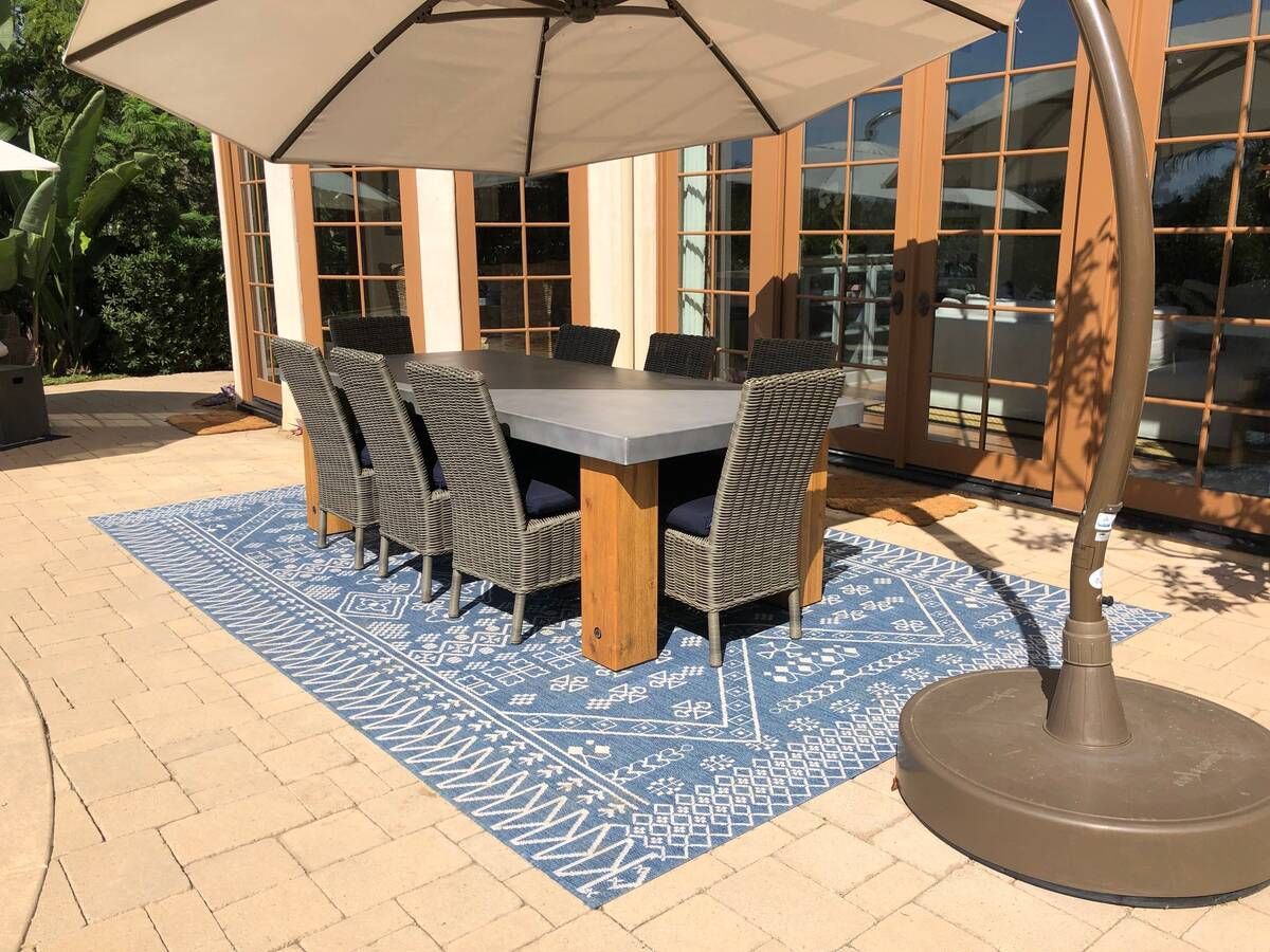 Outdoor dining set: rectangular table, blue rug, gray chairs under a beige umbrella.
