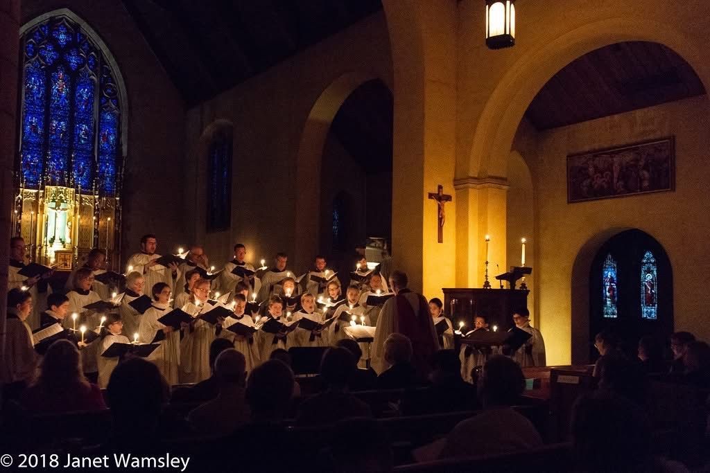 Choir singing in a dimly lit church holding candles, stained glass window.