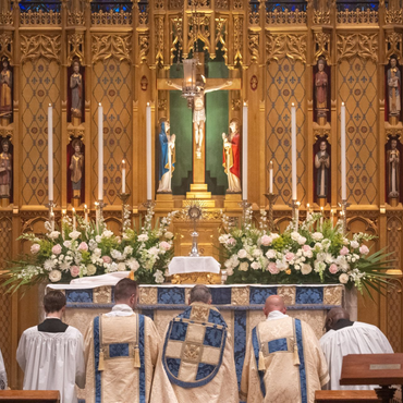 Clergy at the altar facing crucifix during a church service with floral arrangements and statues.