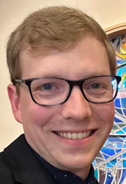 Man with glasses smiles at camera, blond hair, wearing a dark blazer, with a colorful backdrop.
