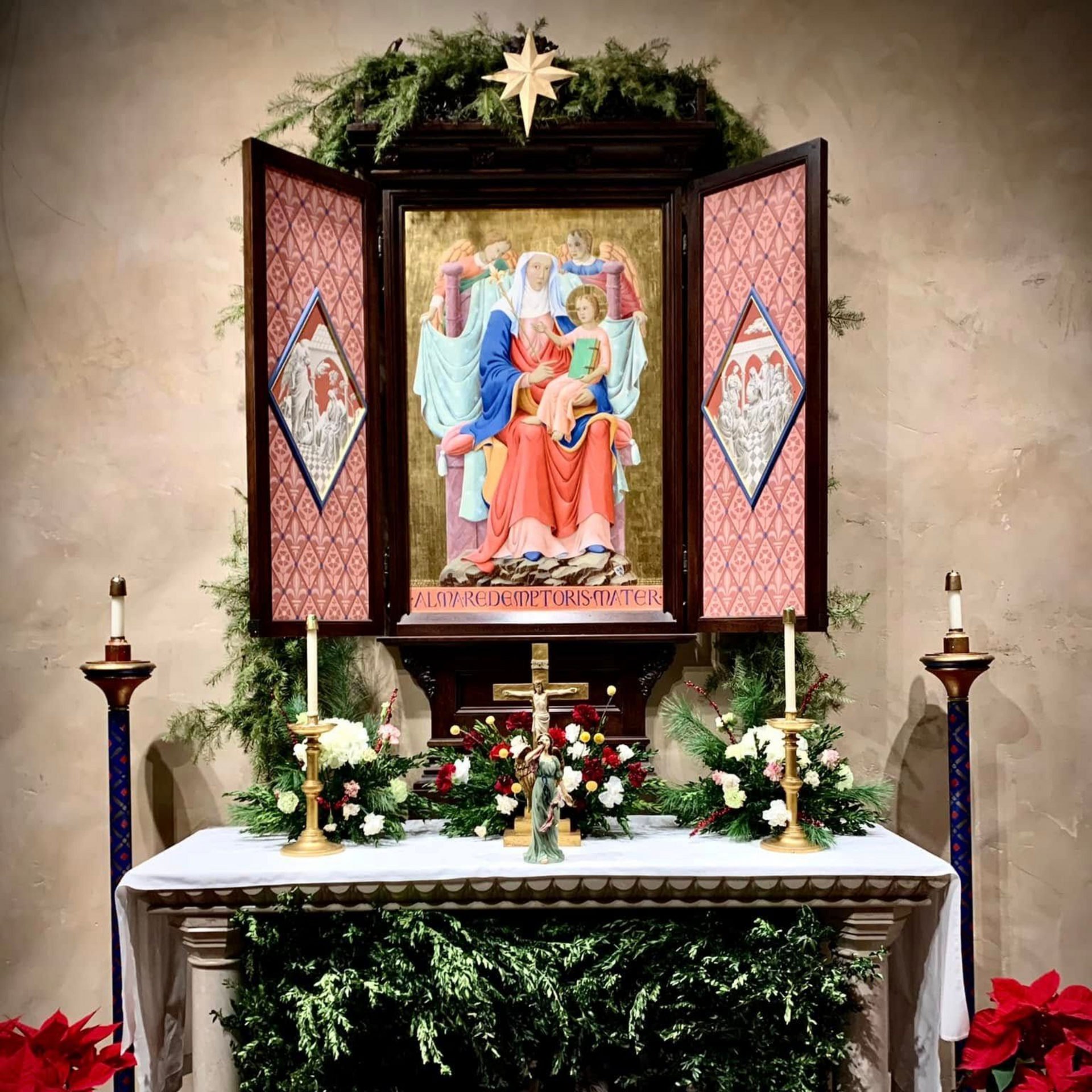 Clergy at the altar facing crucifix during a church service with floral arrangements and statues.