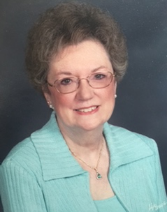 Woman with glasses, wearing a teal jacket and necklace, smiling at the camera.