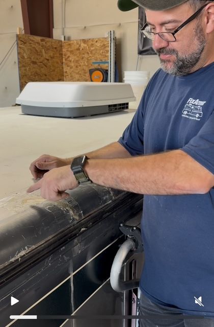 Man in blue shirt works on an RV roof, pointing at a damaged section near a vent in a workshop.