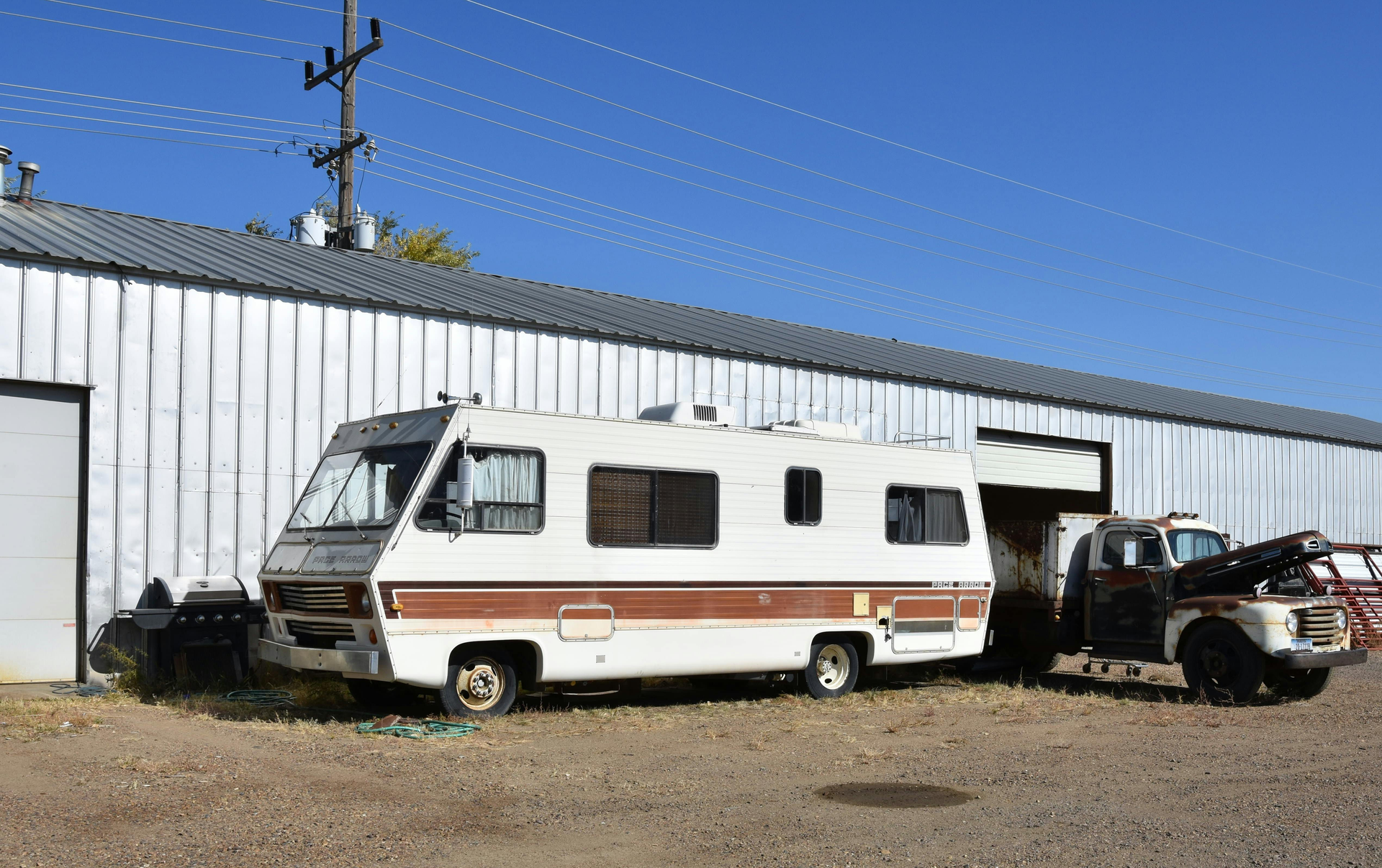 White and brown RV and truck parked in front of a metal building on a sunny day.