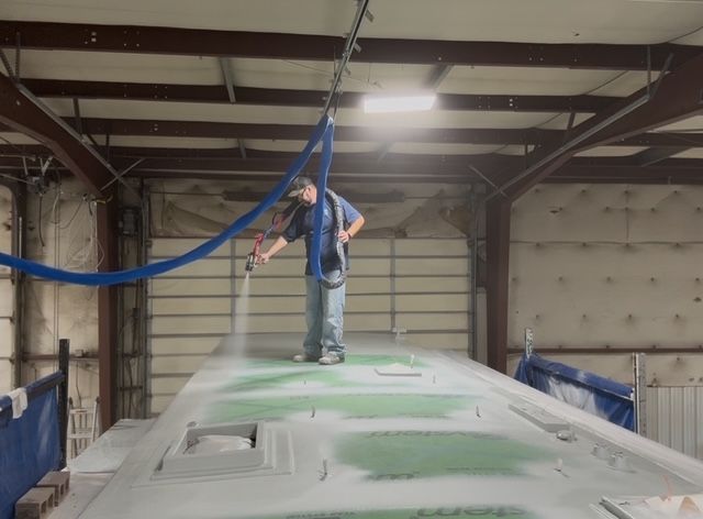 Person sprays paint on a large, flat surface in a warehouse. Blue hose hangs from the ceiling.