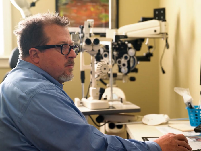 An optometrist in a blue shirt works at a computer in an office with an ophthalmic examination machine in the background.
