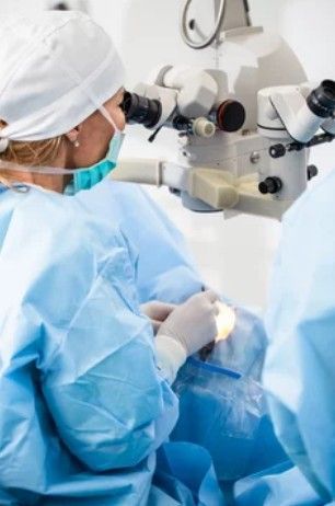 A medical professional in scrubs and a surgical cap performs surgery using a professional-grade clinical microscope.