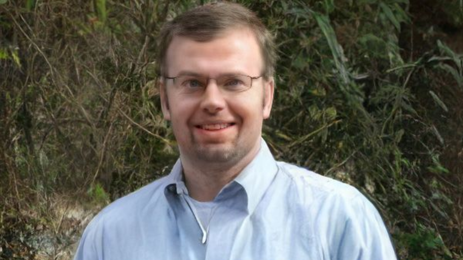 A person with glasses and a light blue collared shirt smiling in front of a blurry outdoor background.