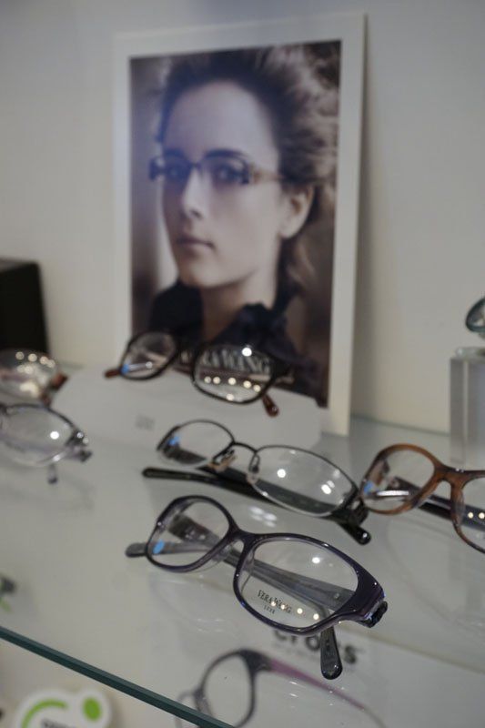 Display of various eyeglass frames on a glass shelf in front of a promotional poster of a person wearing glasses.