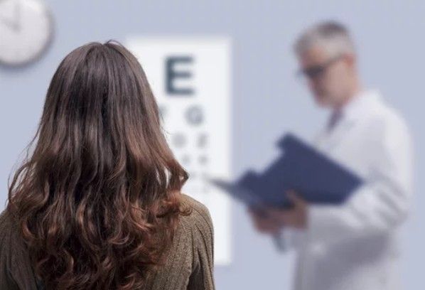 A person faces a blurred eye chart in a doctor’s office as a clinician stands in the background holding a file.