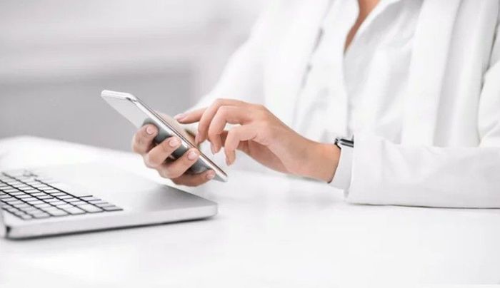 A person in a white blazer sits at a desk with a laptop, using a smartphone.