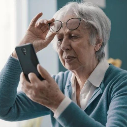 A person wearing glasses holds a smartphone up to read the screen, lifting the glasses to their forehead.
