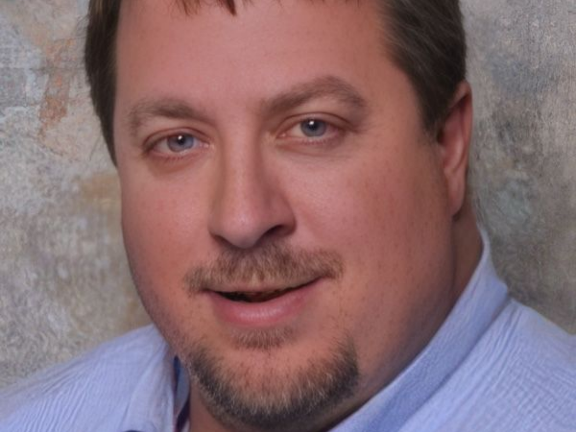 Headshot of a person with a goatee wearing a light blue, collared, button-down shirt against a textured gray background.