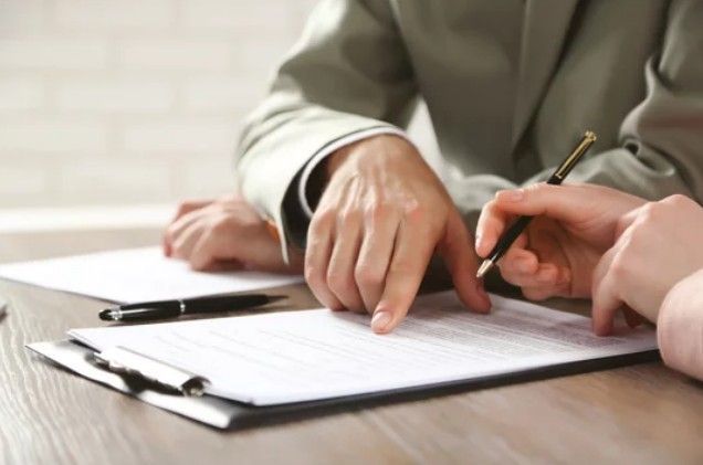 Two people in business attire review a document on a clipboard, with one person pointing and the other holding a pen.