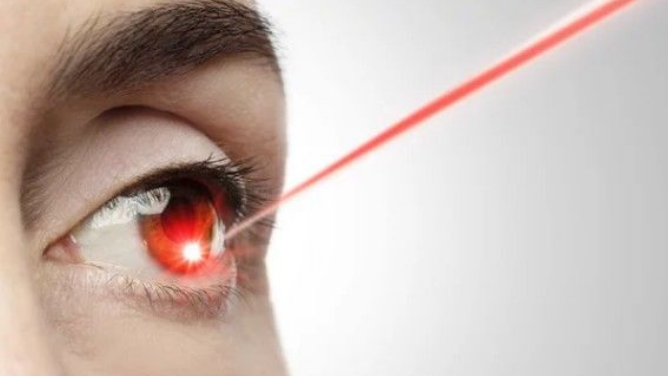 A close-up of an eye with a bright red laser beam shining directly into the pupil against a light grey background.