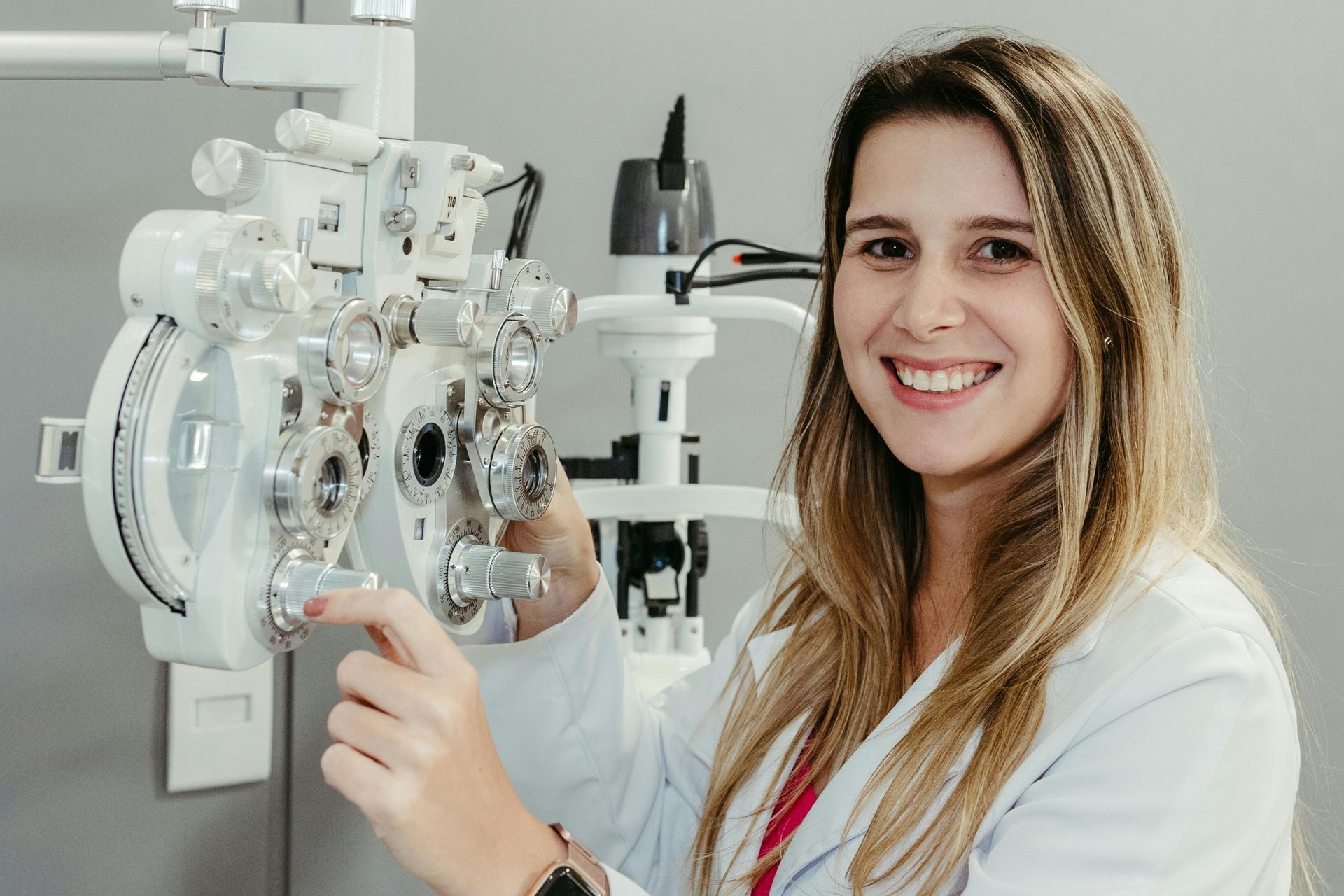A smiling optometrist in a lab coat adjusting the dials on a phoropter in an eye examination clinic.
