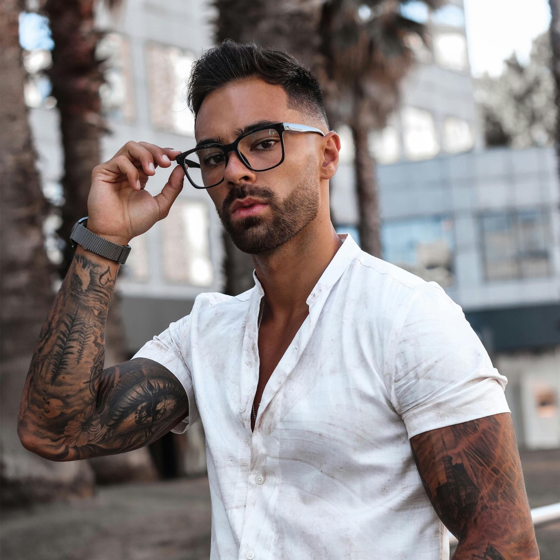 A person with sleeve tattoos wearing glasses and a white button-down shirt poses outdoors near palm trees.