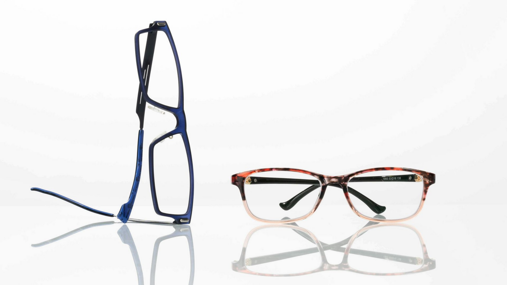 A blue pair of glasses stands vertically next to a pair of tortoise-shell pattern glasses resting on a white surface.