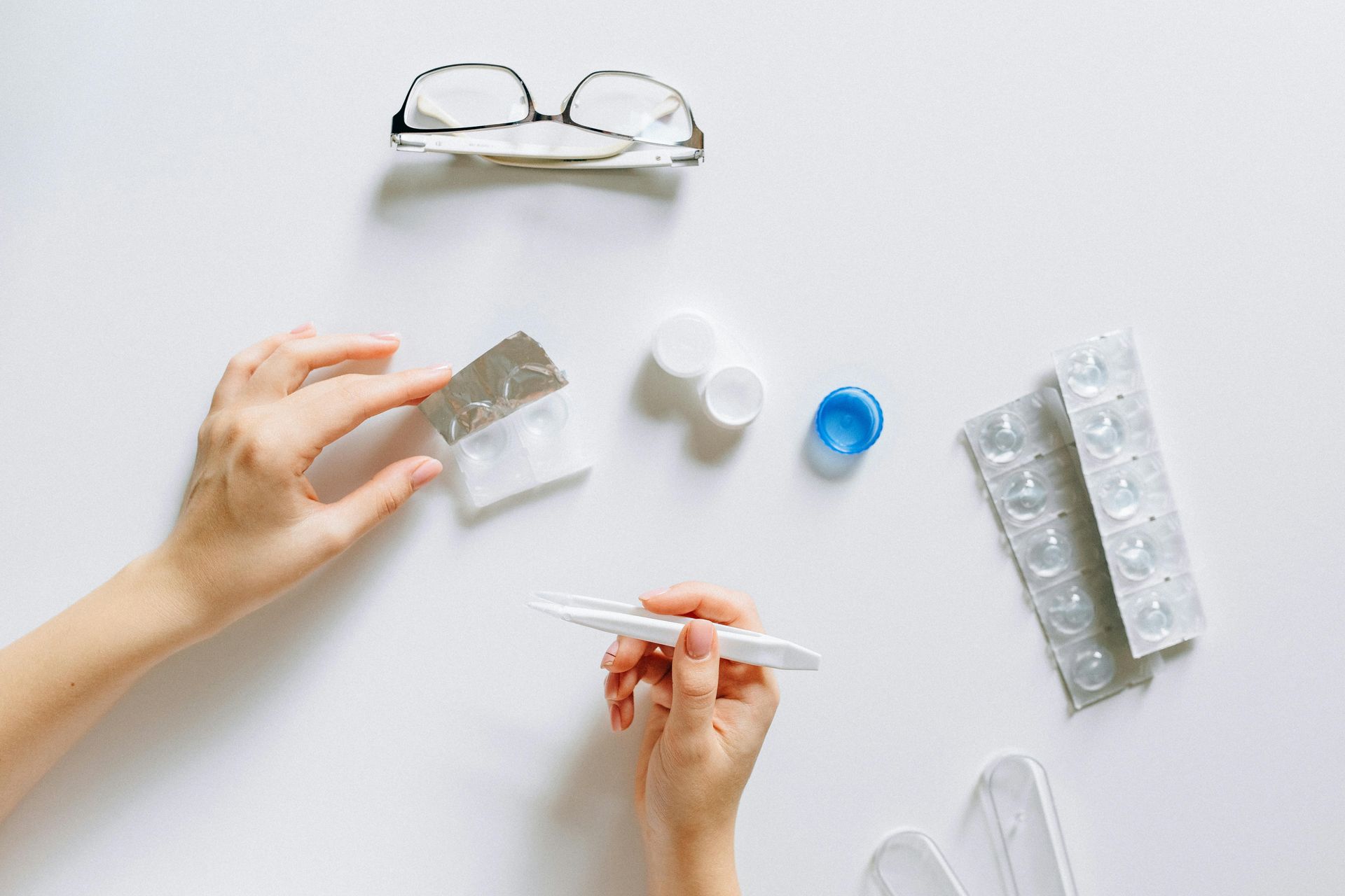 Hands use tweezers and a lens case to handle contact lenses on a white surface with glasses and medication packaging nearby.