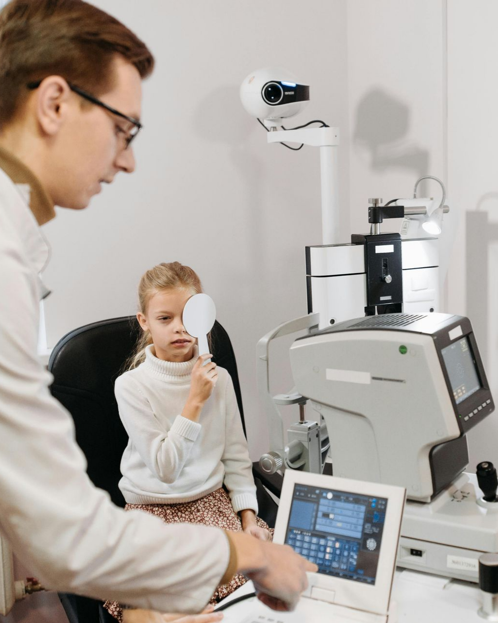 An optometrist points to a digital screen while a patient holds an occluder over one eye during an eye exam.