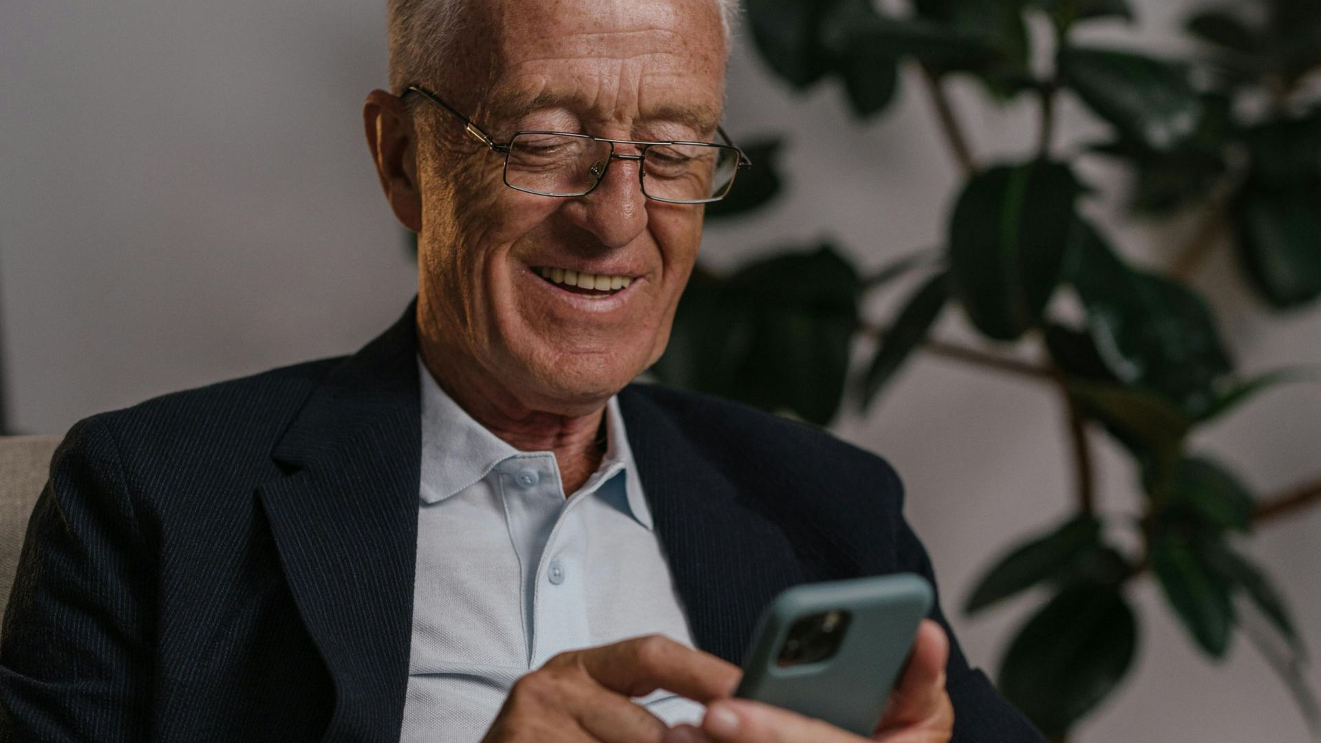 A person wearing glasses and a blazer smiles while looking at a smartphone, seated indoors near a leafy houseplant.