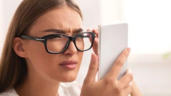 A person wearing black-rimmed glasses squints while holding a smartphone close to their face.