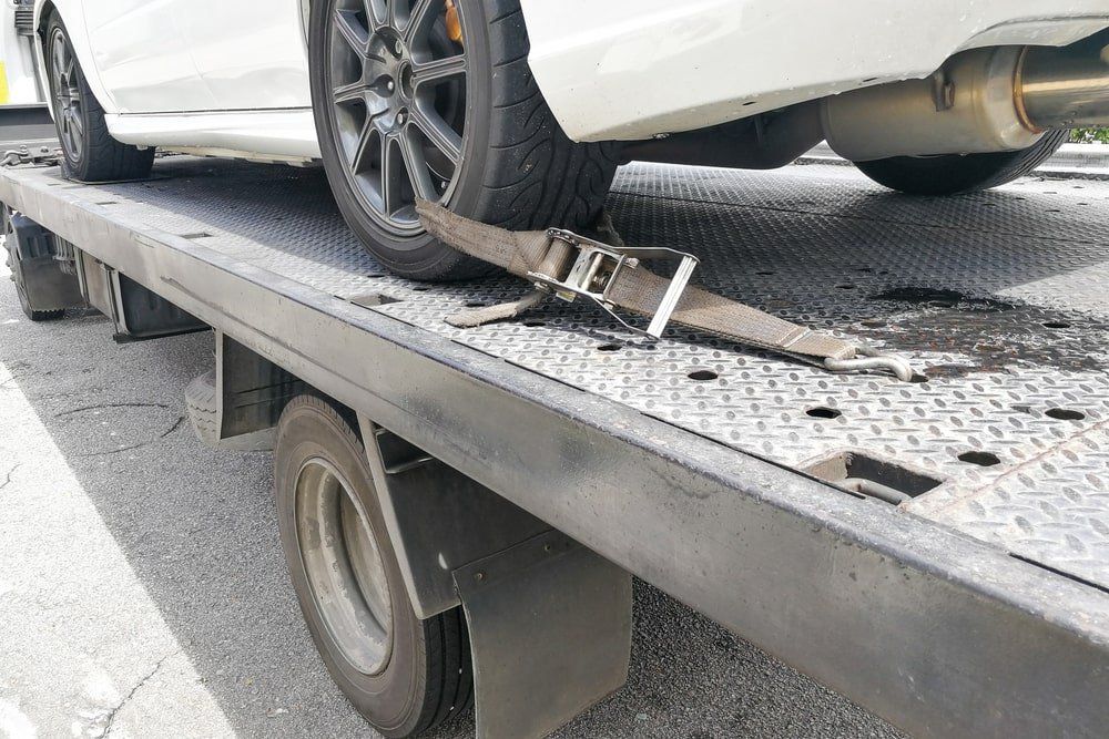 White coupe loaded onto a black flatbed tow truck with ratchet straps connected to the wheels.