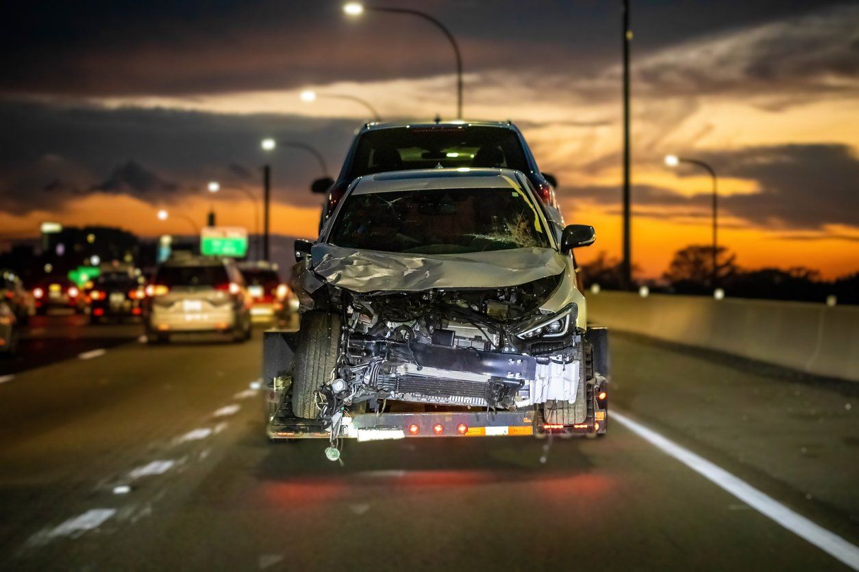 A white car that has been in a car accident being towed on the highway with a sunset background