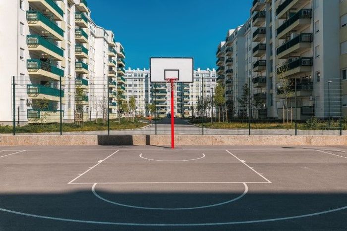 Basketball court with a red hoop, flanked by apartment buildings on a sunny day.
