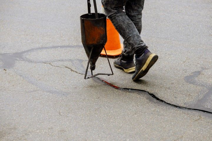 Person filling a crack in asphalt with a tool; orange traffic cone in the background.
