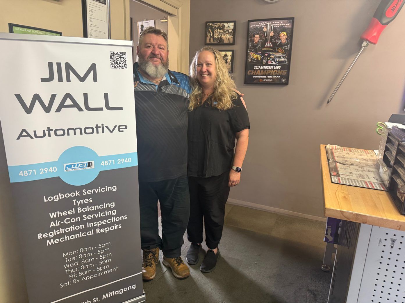 A Man and woman are standing next to their mechanic poster — Jim Wall Automotive in Bowral, NSW
