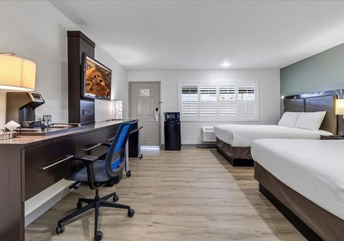 A hotel room with two white beds, a long dark desk with a blue chair, a mounted TV, and a door on a light wood floor.
