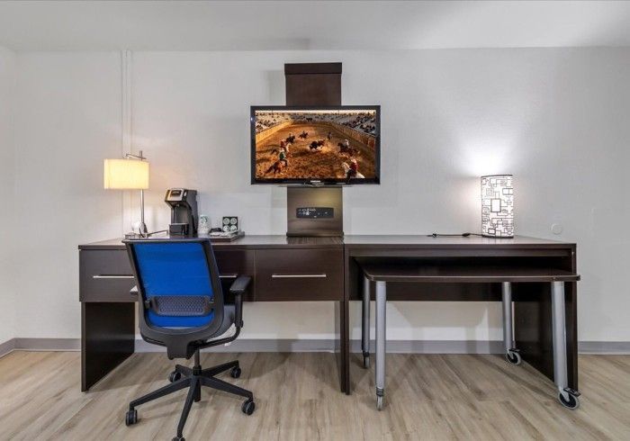 A desk with a blue chair, coffee maker, lamps, and a wall-mounted TV displaying a nature scene in a hotel room.